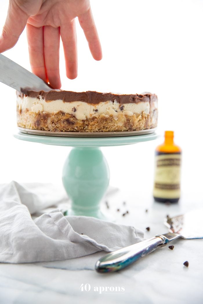 Hand and knife cutting into a vegan no bake cookie dough cheesecake on a cake stand