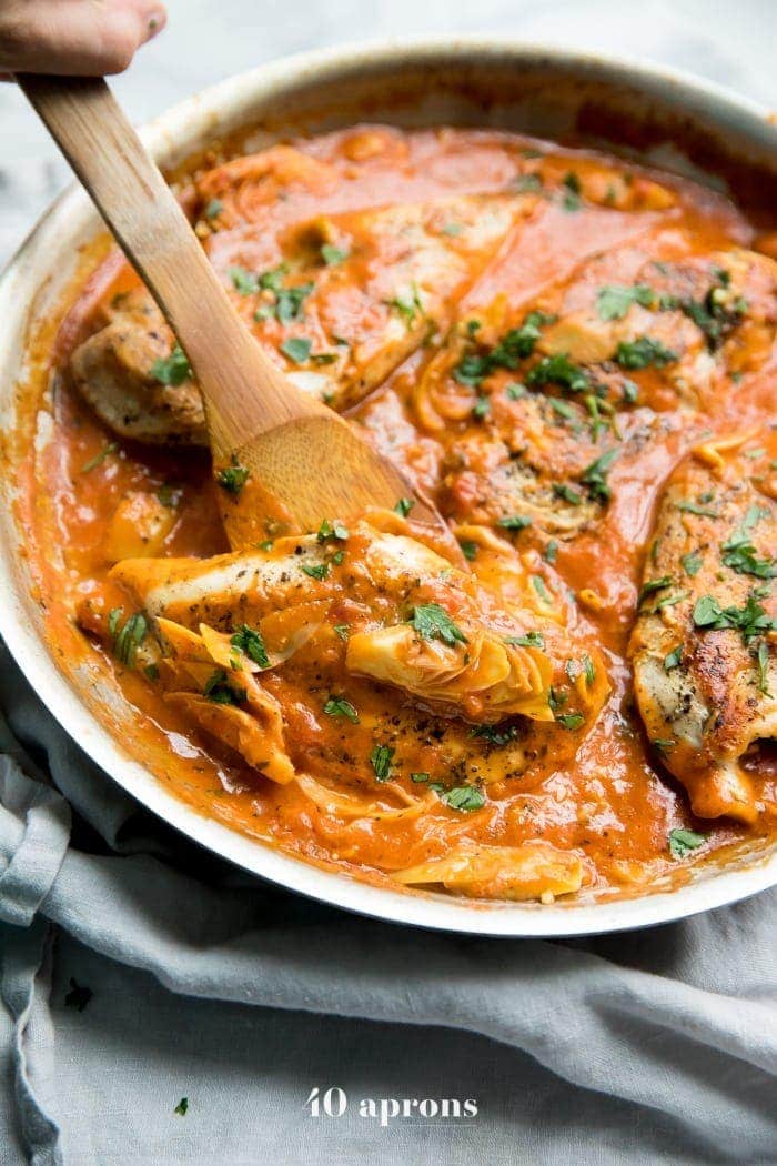Creamy tomato artichoke chicken (Whole30, paleo) in a skillet with a spoon lifting up one chicken piece
