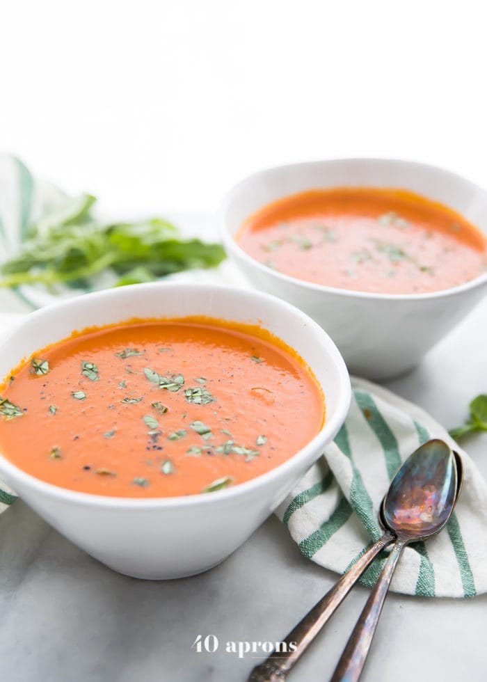 side view of Perfect Whole30 Instant Pot tomato soup (vegan) in two bowls with two spoons and fresh herbs in the background.