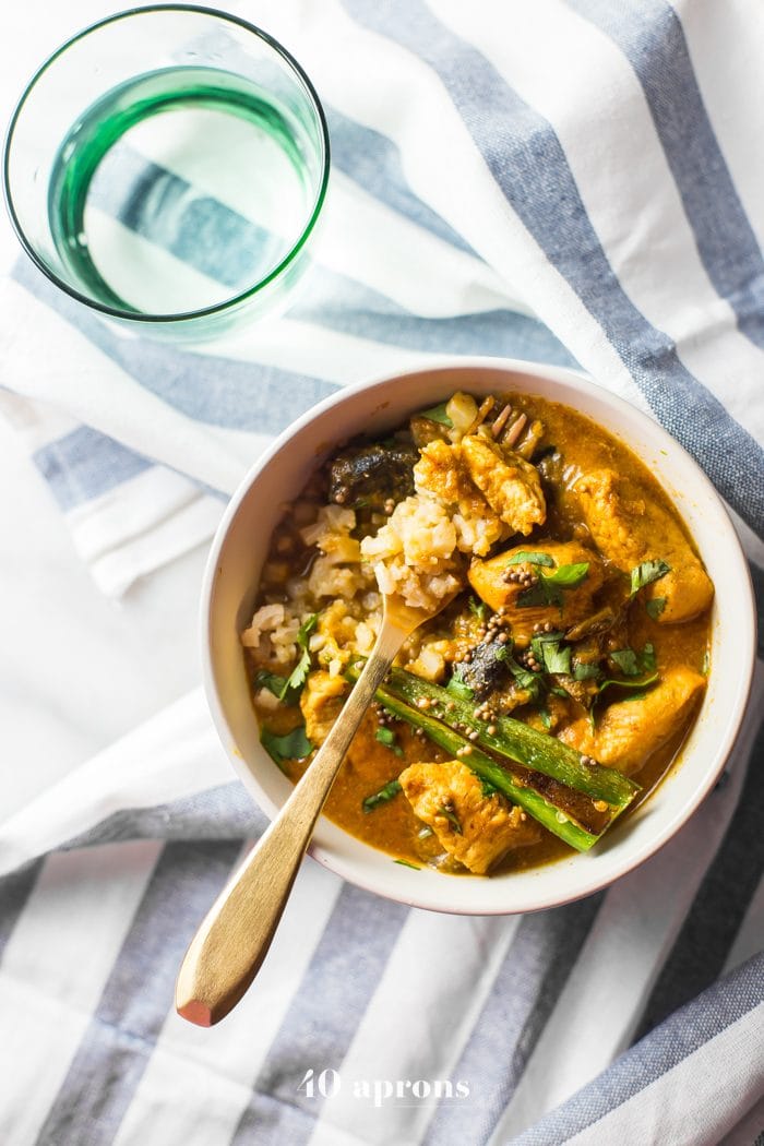 Overhead shot of Whole30 Chicken Curry with cauliflower rice in a white bowl placed on a striped tablecloth with a glass of water on the side.