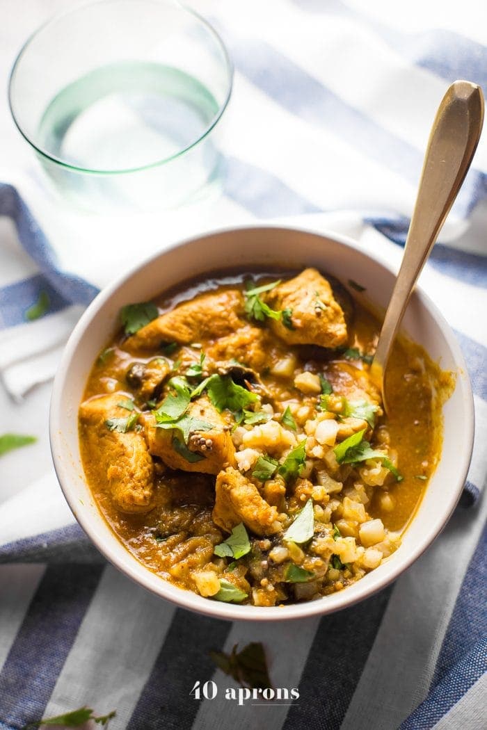 Overhead shot of Whole30 Chicken Curry with a spoon sitting in it, served in a white bowl placed on a striped tablecloth with a glass of water on the side.