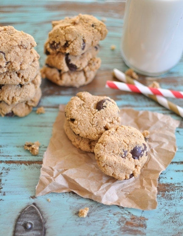 Paleo Chocolate Chip Cookies stacked on top of each other on brown parchment paper with some straws and a glass of milk in the background.