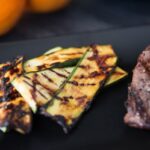 Close-up of slices of orange glazed grilled zucchini crisscross grill marks arranged on a black plate next to a steak.