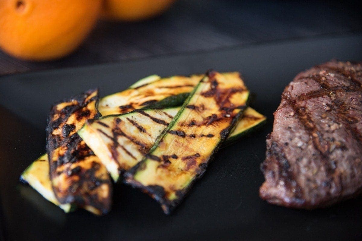 Close-up of slices of orange glazed grilled zucchini crisscross grill marks arranged on a black plate next to a steak.