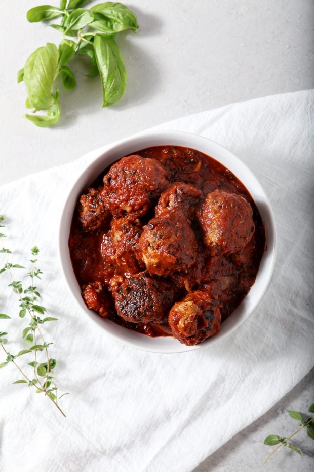 Overhead shot of Whole30 Meatballs in tomato sauce served in a white bowl on a white tablecloth with fresh herbs in the background.