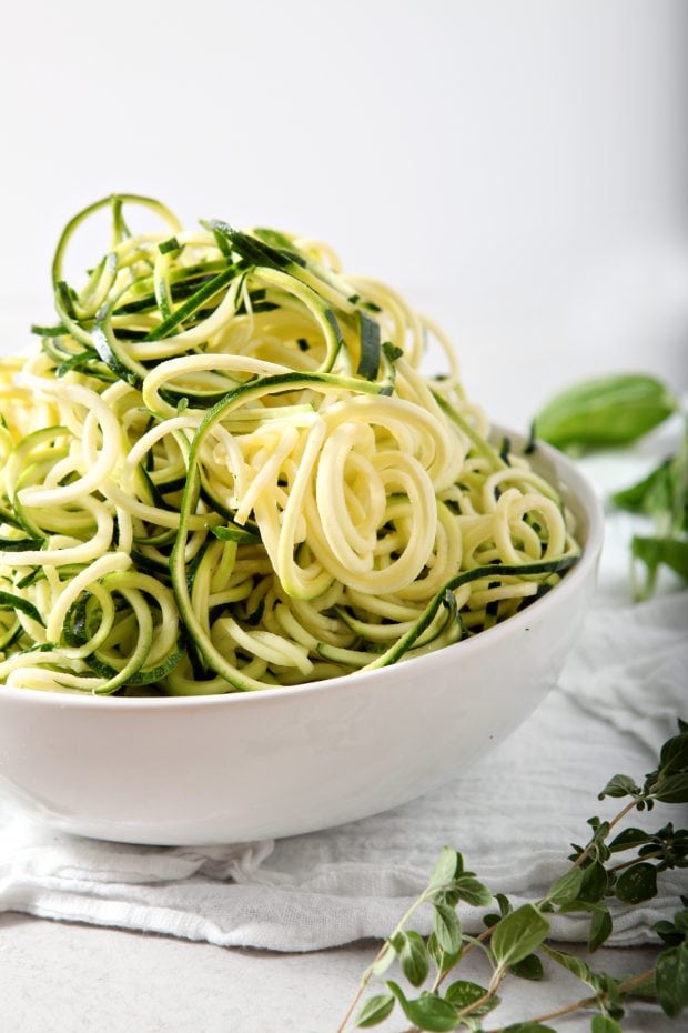 side view of raw zucchini noodles in a white bowl on a white tablecloth with some fresh herbs in the background