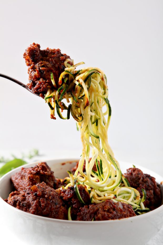 a fork loaded with Whole30 Spaghetti and Meatballs balanced over a white bowl with more zucchini noodles and meatballs.