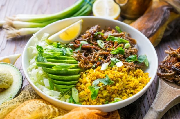 Side view of Instant Pot Brisket Taco Bowl topped with avocado slices, a lemon wedge and chopped green onions placed on a rustic wooden tabletop.