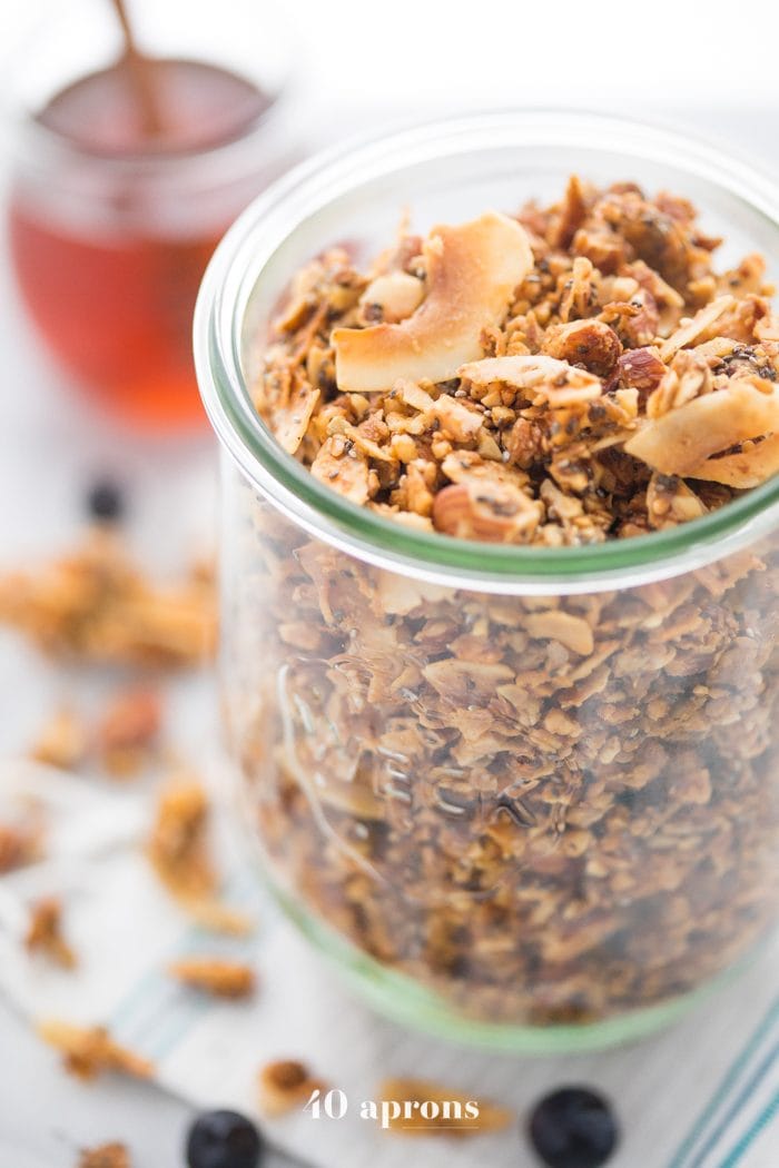 A glass jar of a crunchy paleo granola recipe with a jar of honey in the background