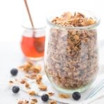 A glass jar of a crunchy paleo granola recipe with a jar of honey in the background