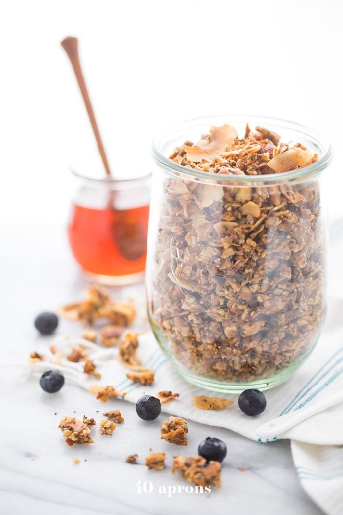 A glass jar of a crunchy paleo granola recipe with a jar of honey in the background