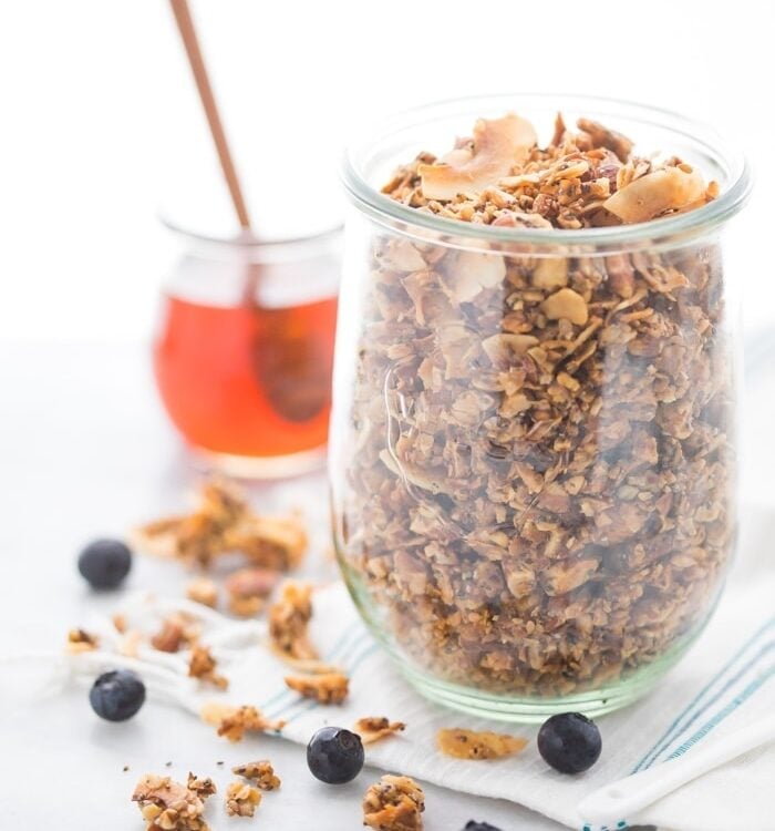 A glass jar of a crunchy paleo granola recipe with a jar of honey in the background