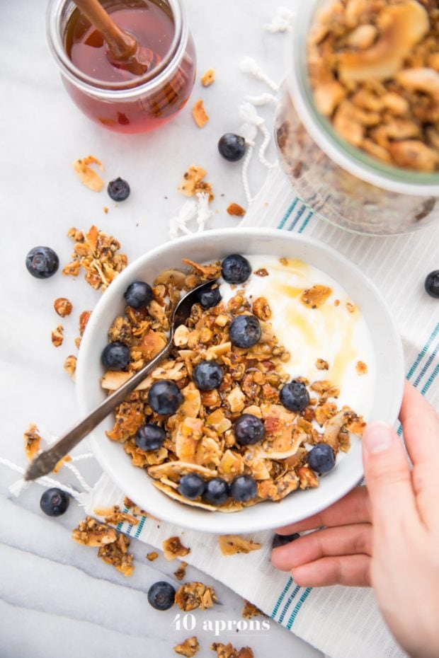 Hand holding a bowl of the crunchy paleo granola recipe over yogurt, topped with blueberries and a drizzle of honey