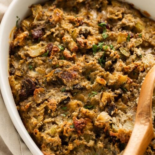 A wooden spoon rests on paleo stuffing in an oval ceramic casserole dish on a holiday table.