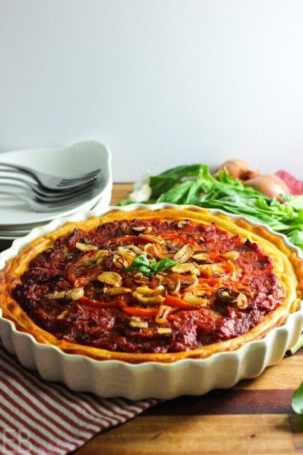 Whole30 Tomato Tart in a white tart dish on a striped kitchen towel with some plates and fresh basil in the background