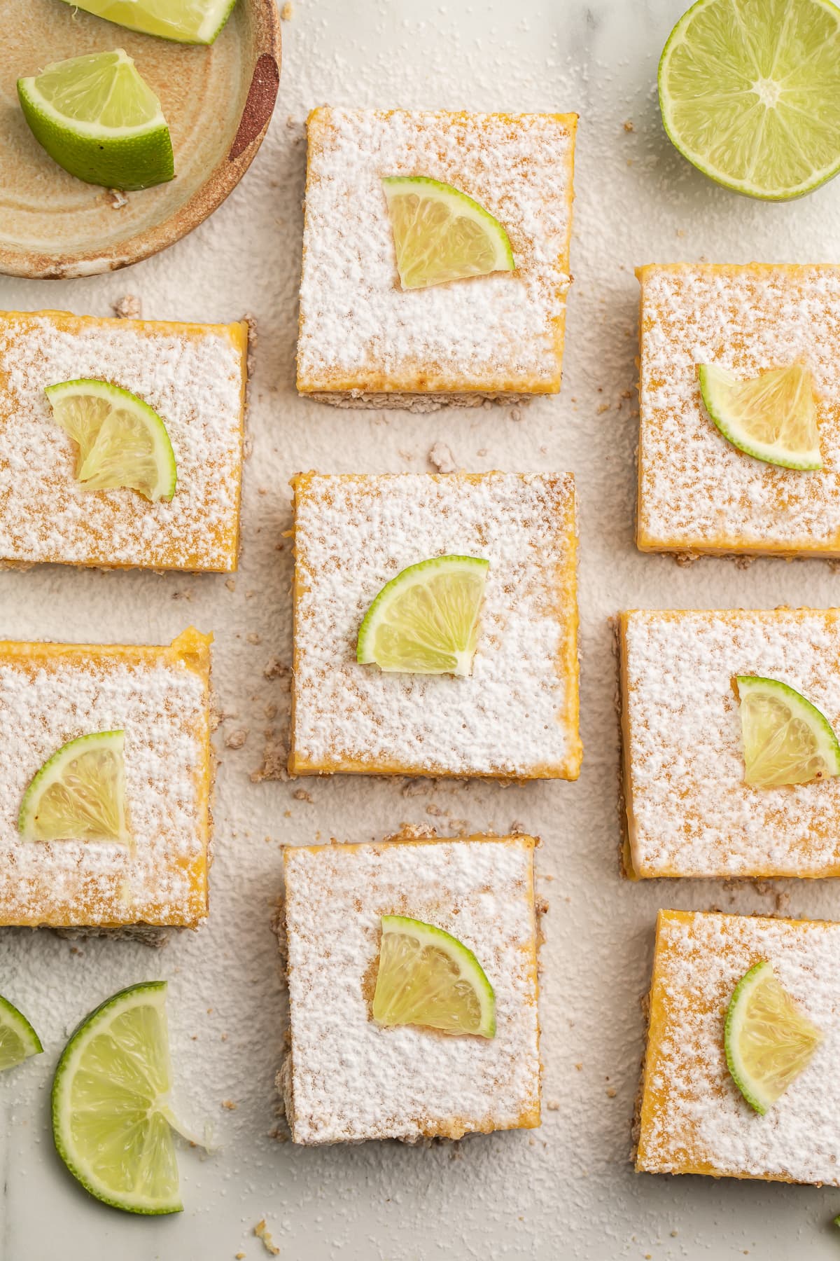 Top down view of 8 paleo lime bars on a table in columns, with powdered sugar and lime wedges.