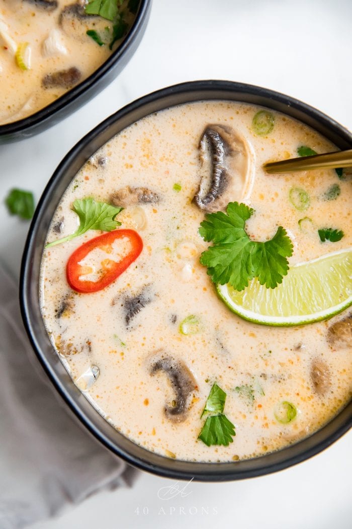 A close up shot of a black bowl of the best tom kha gai Thai chicken coconut soup with limes, lemongrass, chiles, cilantro, and chicken in a coconut broth