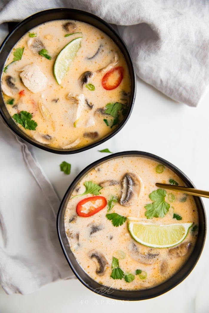 An overhead shot of two black bowls of the best Thai coconut tom kha gai chicken soup with lime, lemongrass, chillies, cilantro and chicken in a coconut broth