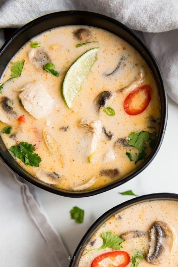 An overhead shot of two black bowls of the best tom kha gai Thai chicken coconut soup with limes, lemongrass, chiles, cilantro, and chicken in a coconut broth