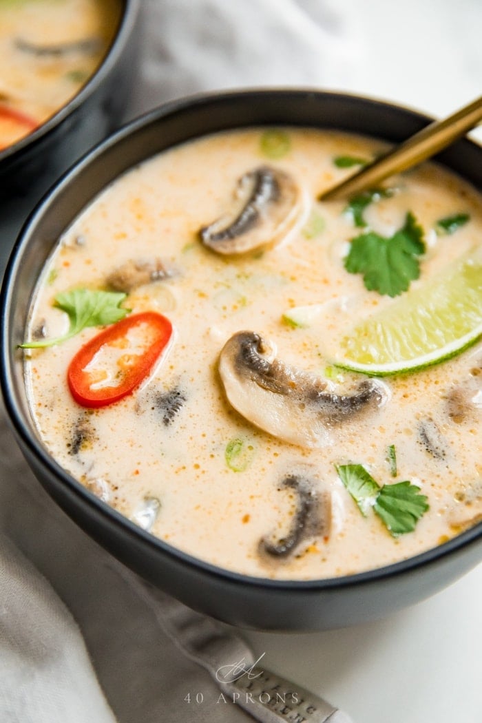 A close up shot of a black bowl of the best Thai coconut tom kha gai chicken soup with lime, lemongrass, chillies, cilantro and chicken in a coconut broth