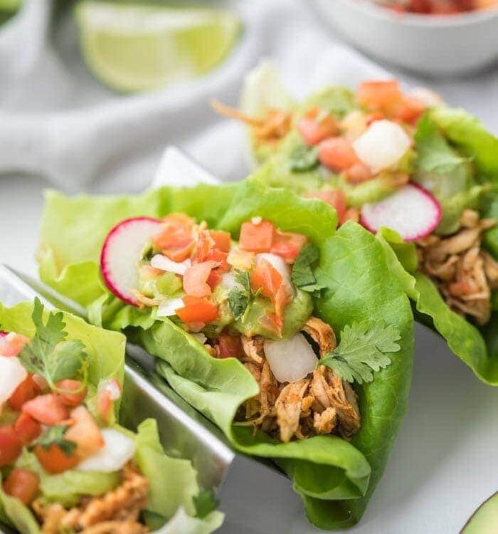 Three paleo chicken tacos in a taco holder served in lettuce wraps topped with avocado crema, pico de gallo, and radishes