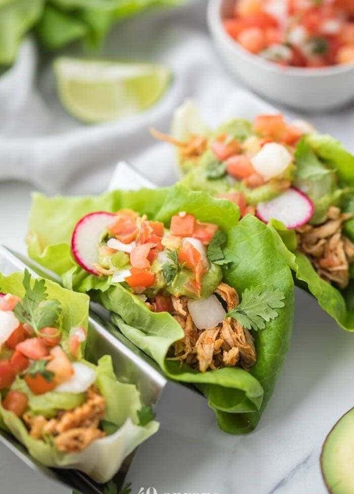 Three paleo chicken tacos in a taco holder served in lettuce wraps topped with avocado crema, pico de gallo, and radishes