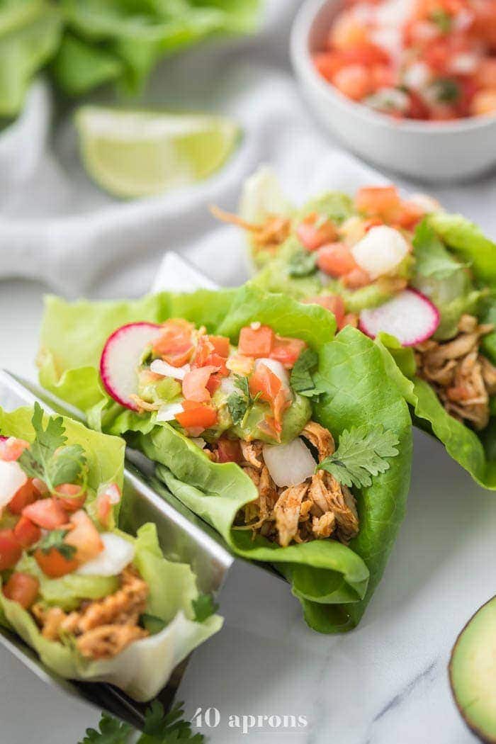 Three paleo chicken tacos in a taco holder served in lettuce wraps topped with avocado crema, pico de gallo, and radishes