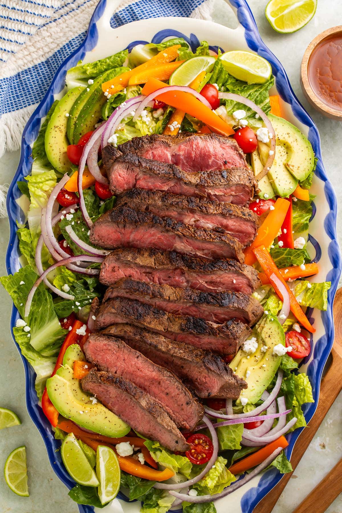 Fajita steak salad arranged on a blue and white platter with lime wedges and slices of avocado.