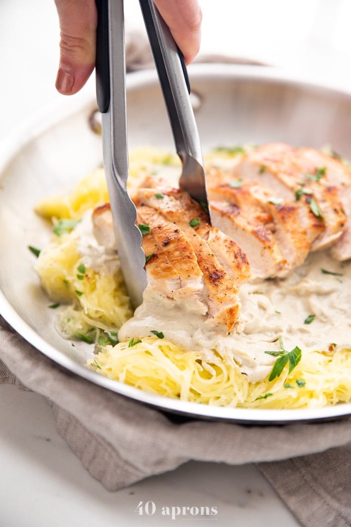 Healthy Whole30 chicken alfredo with spaghetti squash in a skillet topped with parsley and tongs lifting up