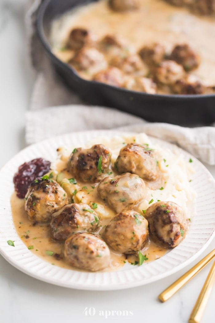 A plate of paleo Swedish meatballs over mashed potatoes with Swedish meatballs sauce and a side of raspberry jam with skillet in the background