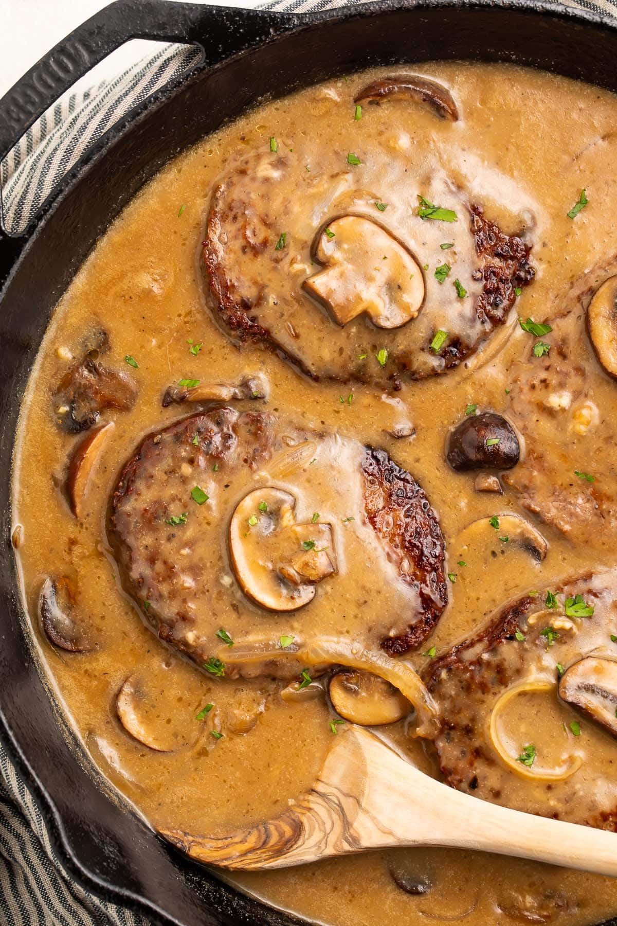 Four Whole30 salisbury steaks in a cast-iron skillet with plenty of salisbury sauce, mushrooms, and a wooden spoon.