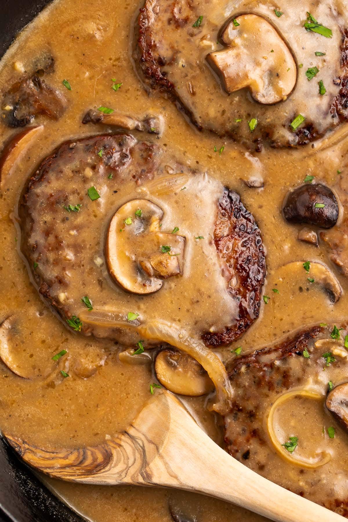 Whole30 salisbury steak in a cast-iron skillet with plenty of brown gravy, mushrooms, and a wooden spoon.