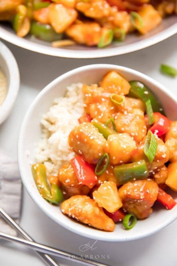 Healthy sweet and sour chicken with peppers over cauliflower rice in a white bowl with sesame seeds and chopsticks to the side