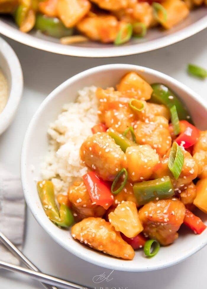 Healthy sweet and sour chicken with peppers over cauliflower rice in a white bowl with sesame seeds and chopsticks to the side