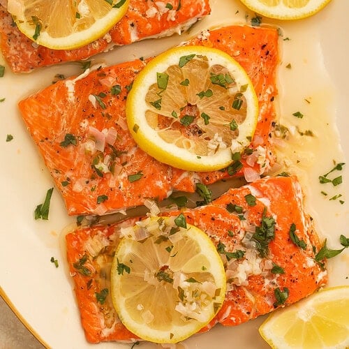 Fillets of salmon in a lemon butter sauce on a cream serving platter. The salmon is topped with lemon slices and chopped herbs.