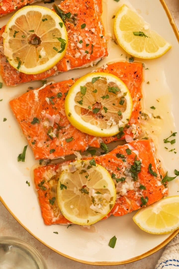 Fillets of salmon in a lemon butter sauce on a cream serving platter. The salmon is topped with lemon slices and chopped herbs.