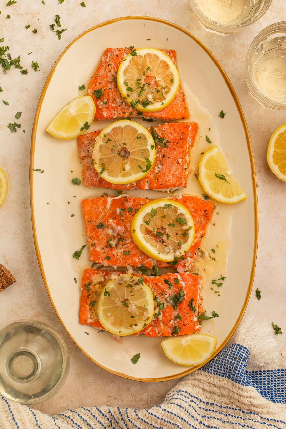 Platter of cooked salmon fillets in a buttery sauce topped with sliced lemons and chopped herbs.