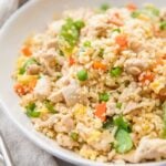 A white bowl of cauliflower fried rice with chicken, peas, carrots with a grey linen and silver chopsticks to the side