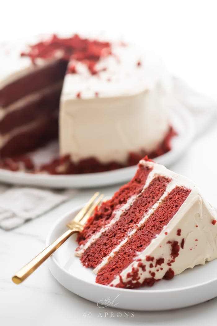 Slice of three layer gluten free red velvet cake with paleo cream cheese frosting on a white plate with whole cake in background