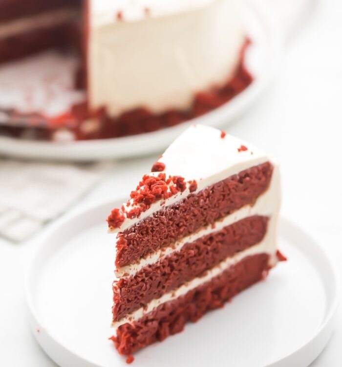 Slice of gluten free red velvet layer cake with paleo cream cheese frosting on a white plate with whole cake in background