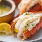 Broiled lobster tails butterflied on a plate with parsley and a lemon wedge next to a wooden bowl of garlic butter dipping sauce