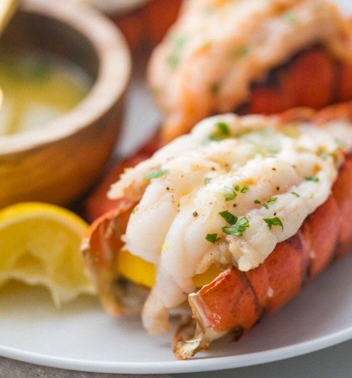 Broiled lobster tails butterflied on a plate with parsley and a lemon wedge next to a wooden bowl of garlic butter dipping sauce