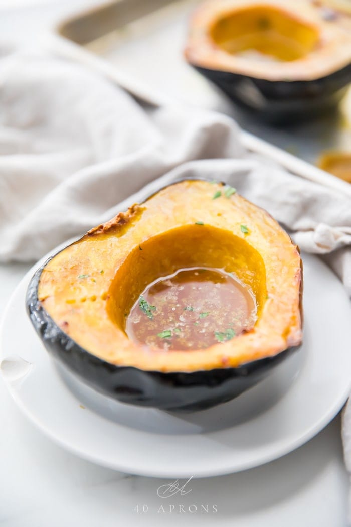 A roasted acorn squash half filled with melted maple butter on a white plate and a grey linen behind