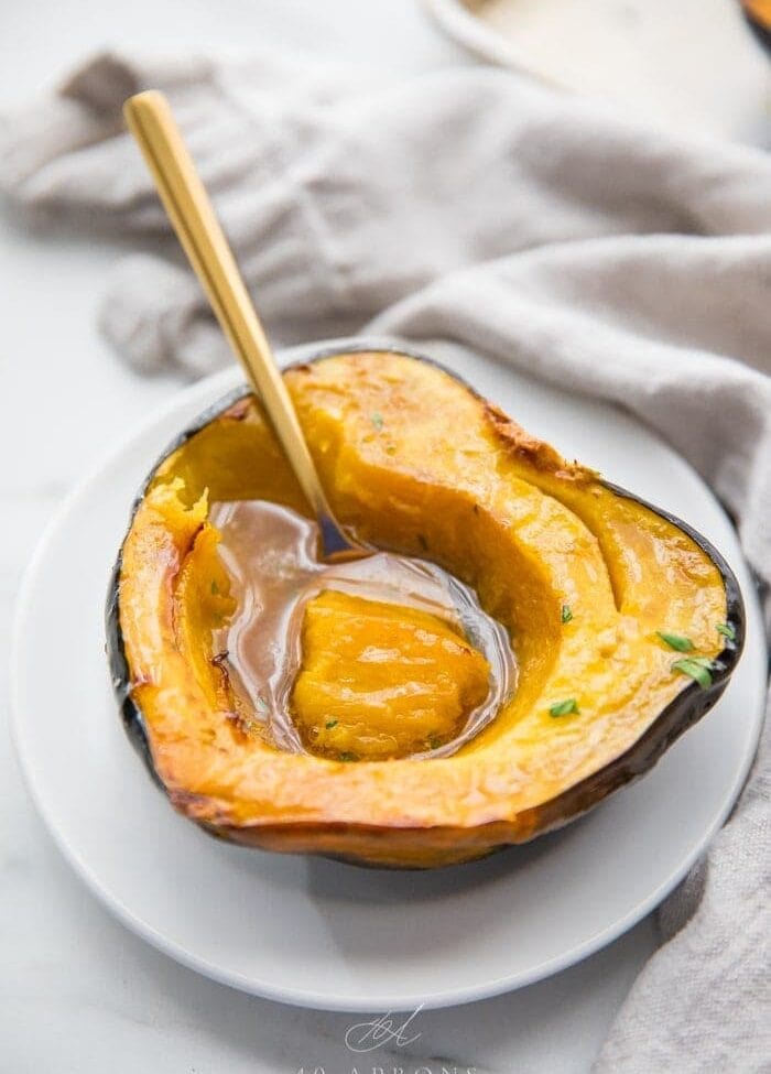 A roasted acorn squash half filled with melted maple butter on a white plate with a gold spoon scooping out a bite