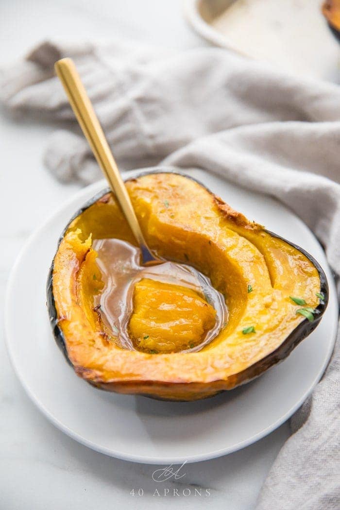 A roasted acorn squash half filled with melted maple butter on a white plate with a gold spoon scooping out a bite