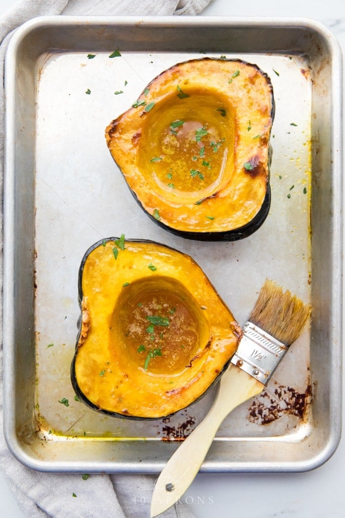 Two roasted acorn squash halves with melted maple butter in the centers on a baking sheet with a pastry brush