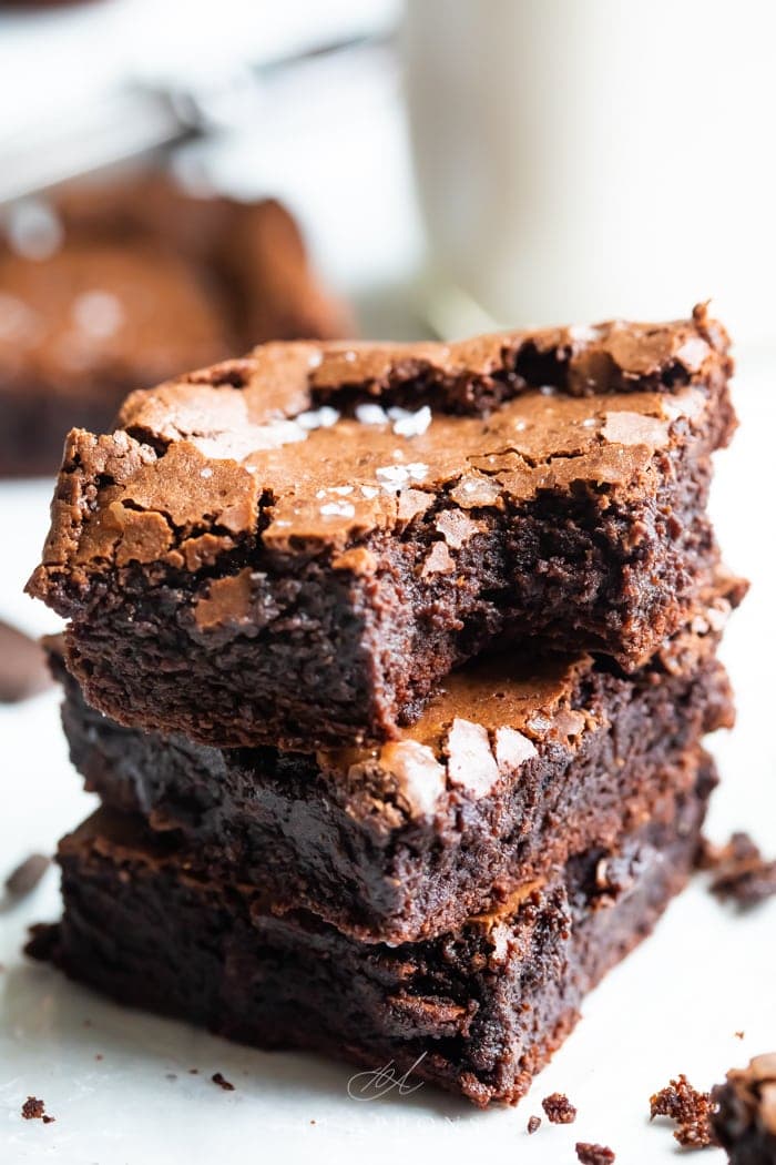 A stack of three gluten free, paleo brownies showing how fudgy and rich they are, with a crackly top and flake sea salt on top and a bite out of one