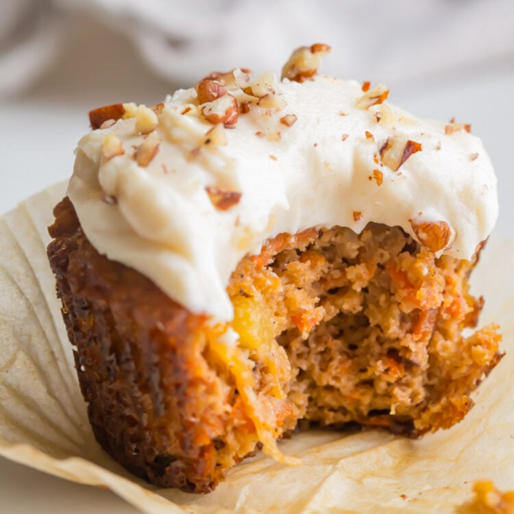 A paleo carrot cake cupcake topped with cream cheese frosting and chopped pecans. A bite is missing from the cupcake.