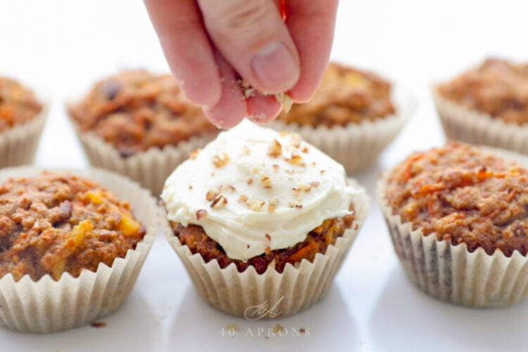 A white woman's hand sprinkling finely chopped pecans onto the top of frosted carrot cake cupcakes.