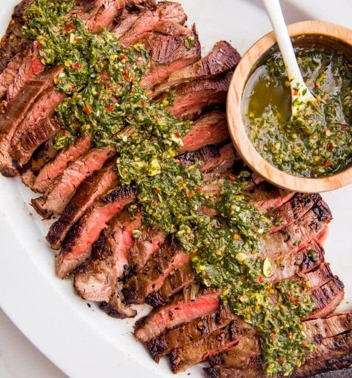 Sliced medium rare flank steak on a plate with lots of fresh chimichurri sauce on top